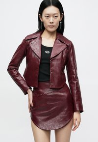 Vrouw die een bordeauxrode cropped jas van nepleer en een bijpassende minirok draagt over een zwart shirt met logo, staand tegen een effen achtergrond.