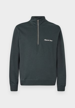 Mørkegrå hettegenser med høy krage og halv glidelås. Har hvit brodert tekst som sier "Originals Studio" på venstre bryst.