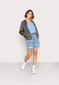 Casaco bomber floral com fundo preto, blusa azul de decote em V, calções de ganga desgastados e ténis brancos. Look casual, com um ajuste relaxado.