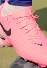 Rózsaszín Nike foci cipő, textúrált felülettel, fekete márkajelzéssel, lapos fűzőkkel és áramvonalas kialakítással. Egy focilabdán helyezkedik el.