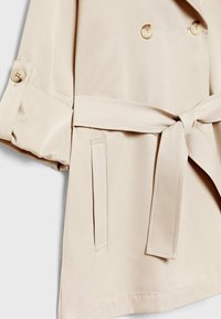 Beige dubbelbreasted trenchcoat gemaakt van gladde stof. Voorzien van een ceintuur die kan worden gestrikt, twee zijzakken en knoopmanchetten.