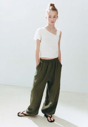 Jeune personne avec les cheveux en chignon portant un haut blanc asymétrique à manches courtes, un pantalon ample vert olive et des tongs noires, debout à l'intérieur.