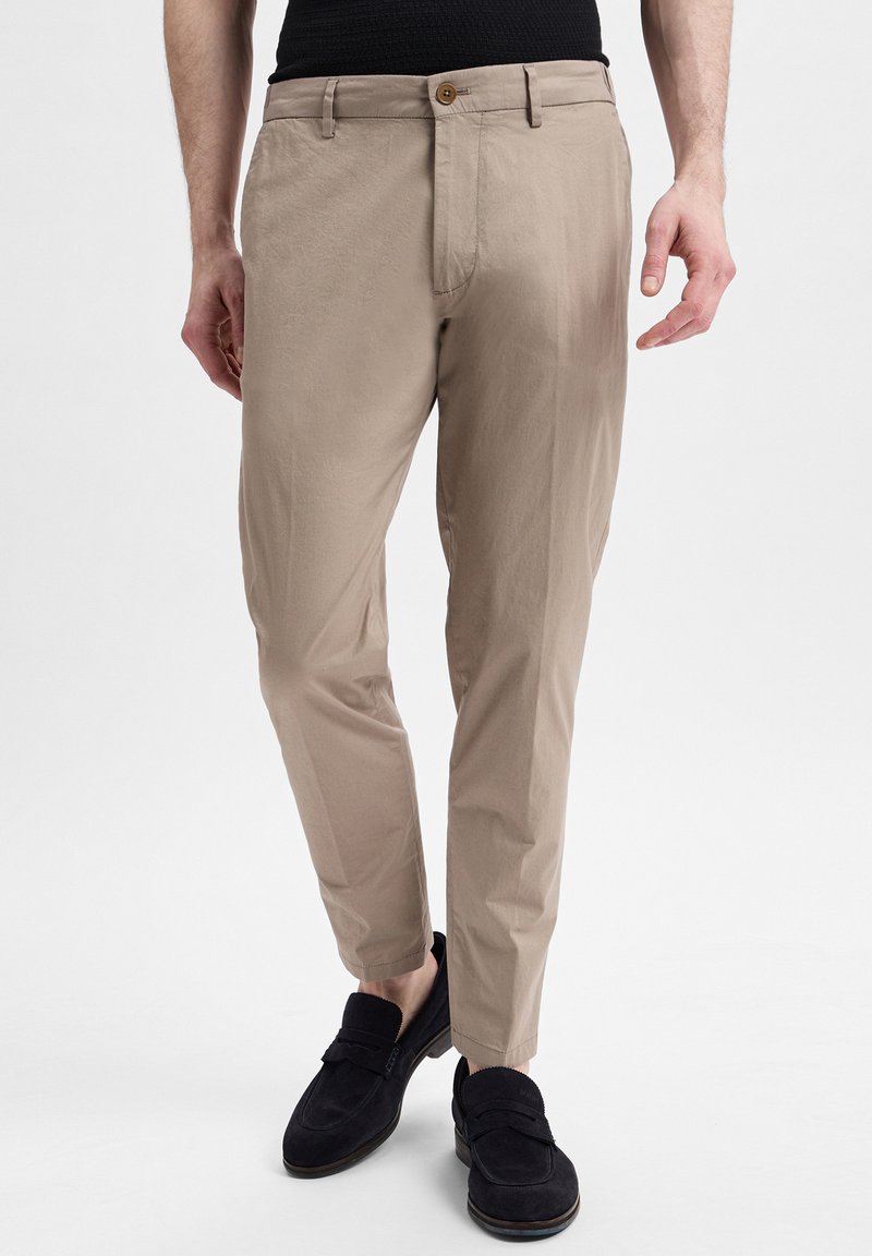 Beige tapered trousers with crease details, front pockets, and a single button closure, paired with dark loafers. Smooth fabric texture.