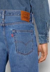 Jean en denim bleu à coupe classique, avec un patch en cuir marron, un logo sur une étiquette rouge, et une couture arcuée sur la poche arrière.