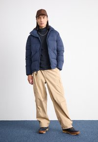 Marineblå pufferjakke med glidelås, hette og sidelommer, sammen med beige cargo-bukser og mørke sko, mot en nøytral bakgrunn.