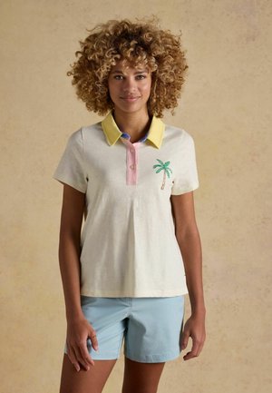 Kobieta o kręconych włosach, ubrana w koszulkę polo z krótkim rękawem z żółtym kołnierzykiem i haftem palmy, w połączeniu z jasnoniebieskimi szortami.
