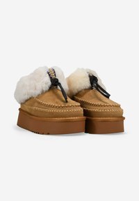 Zapatillas de plataforma de ante marrón con un grueso forro de piel blanca, costuras decorativas y lazos negros sobre un fondo blanco.