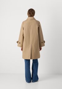 Khakifarbener Trenchcoat mit geradem Schnitt, ausgestattet mit einem Kragen und weiten Ärmeln, akzentuiert durch dunkle Knöpfe. Kombiniert mit blauen Schlagjeans.