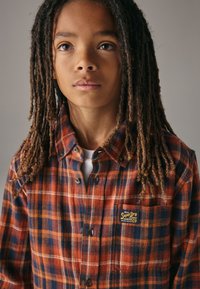 Rød og blå ternet flannel-skjorte med krave, knaplukning og brystlomme med "SuperDry"-label. Struktureret stof.