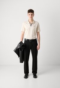 Hellbeige Polo mit einem gestreiften Kragen, schwarzem Gürtel, schwarzen Hosen und einer über dem Arm getragenen Lederjacke. Schwarze Schuhe runden das Outfit ab.