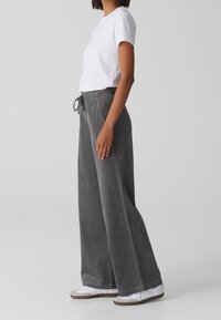 Pantalon large en velours côtelé gris avec taille élastique et cordon de serrage. Associé à un t-shirt blanc uni et des chaussures légères.