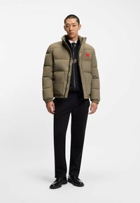 Giacca a vento in verde oliva con un design corto, texture trapuntata e patch con logo rosso. Indossata sopra un top scuro e pantaloni neri.