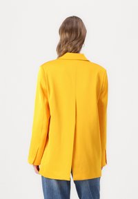 Gul blazer med skreddersydd design, notklapper, lange ermer, og en enkel bakventil, kombinert med blå jeans. Glatt tekstur.
