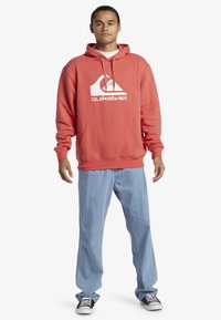 Červená mikina s kapucňou a bielym logom Quiksilver, v kombinácii s svetlomodrými denimovými džínami a čierno-bielymi teniskami. Neformálny strih.