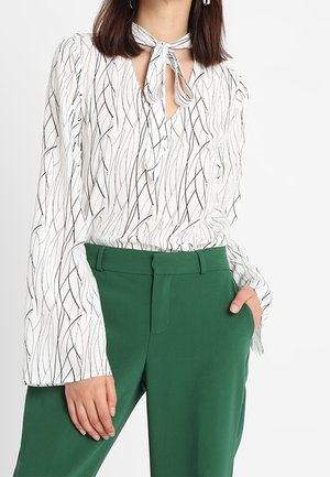 Kobieta ubrana w białą bluzkę z czarnymi falistymi liniami i wiązanym kołnierzykiem, w połączeniu z wysokim stanem zielonych spodni, z ręką w kieszeni.