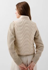 Gilet beige trapuntato con motivi a onda; maniche in maglia di un beige più chiaro; polsini a costine; logo "SCALPERS" sul colletto.