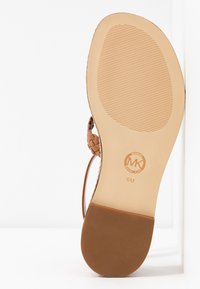 Sandale tressée marron avec une semelle en caoutchouc texturée, présentant une finition lisse et un logo embossé. Le talon est plat, offrant un design ouvert.