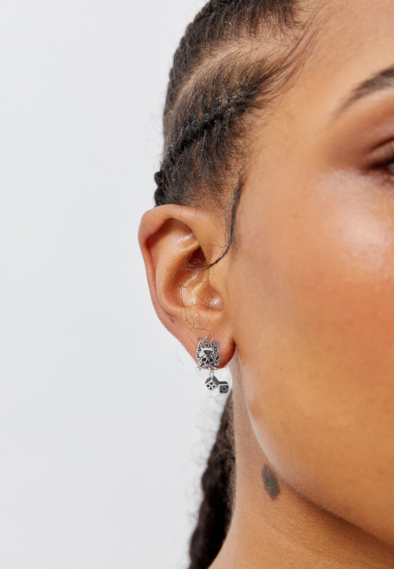 Silver earrings feature a flame and skull design with hanging dice accents. The texture appears smooth, with a shiny metallic finish.