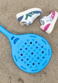 Pala azul con un diseño perforado se encuentra sobre la arena, junto a dos pares de zapatos para niños: uno en azul, blanco y verde; otro en blanco con acentos rosas.