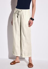 Ljust beige, linnebyxor med vida ben, elastisk midja och dragsko. Har uppvikta fållar och är kombinerade med bruna slidesandaler.