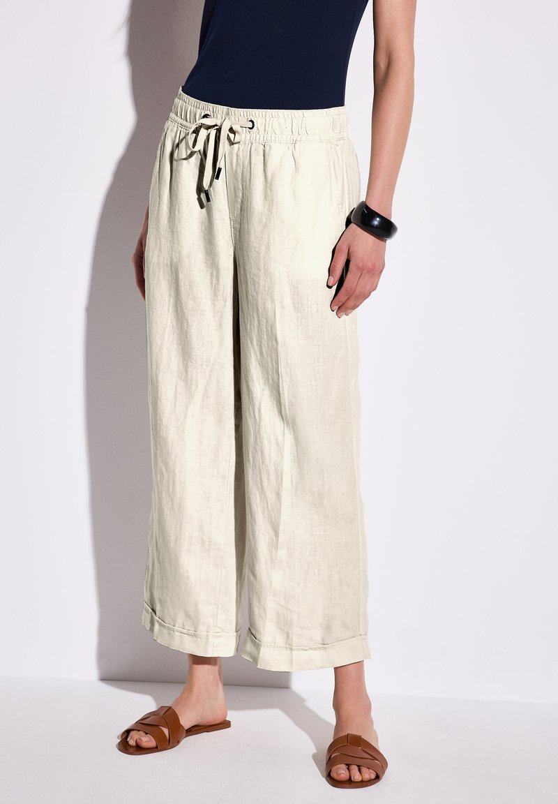 Ljust beige, linnebyxor med vida ben, elastisk midja och dragsko. Har uppvikta fållar och är kombinerade med bruna slidesandaler.