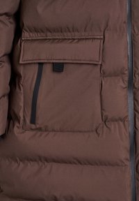 Braune gesteppte Jacke mit Tasche und Klappe sowie vertikalem schwarzem Reißverschlussdetail neben einem schwarzen Reißverschluss, der vorne an der Jacke verläuft.
