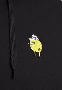 Zwarte hoodie met een klein, geel geborduurd figuurtje dat lijkt op een citroen, met een witte pet waarop "LEMON" staat.