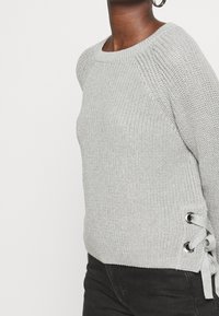 Jasnoszary sweter z dzianiny z okrągłym dekoltem, prążkowanymi krawędziami oraz bocznym sznurowaniem z metalowymi oczkami. Teksturowana tkanina o luźnym kroju.