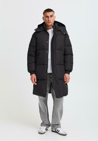 Čierna nafukovacia bunda s kapucňou, s boxovým strihom, prešitým textúrou, zapínaním na cvoky a ribovanými manžetami. Nosená na bielom tričku.