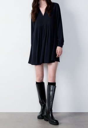 Femme portant une mini-robe noire ample à manches longues associée à des bottes en cuir noires montantes, debout contre un mur clair uni.