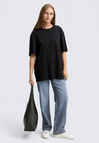 Zwart oversized T-shirt met een ronde hals, korte mouwen en subtiele glinsterdetails, gecombineerd met lichtblauwe jeans en een zwarte draagtas.