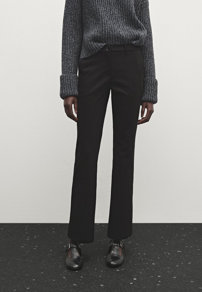 Personne portant un pantalon noir droit, un pull gris côtelé avec les poignets retroussés et des chaussures en cuir noir, debout sur un sol tacheté.