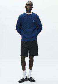 Pull bleu tricoté avec un motif en losange texturé, associé à un short noir, des chaussettes blanches et des mocassins noirs. Tenue simple et minimaliste.