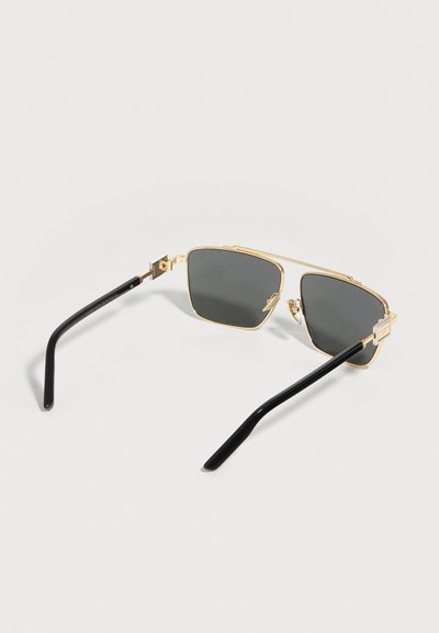 Γυαλιά ηλίου με χρυσό σκελετό, σκοτεινούς ορθογώνιους φακούς και μαύρα γυαλιά, τοποθετημένα πάνω σε φωτεινή επιφάνεια.