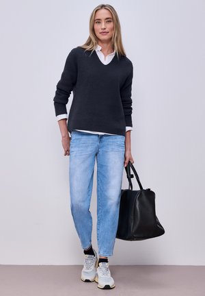 Žena v černém svetru, bílé košili, světle modrých džínách a bílých teniskách, držící velkou černou kabelku, stojící před jednoduchým světlým pozadím.