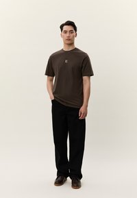 Man står med händerna i fickorna iförd en brun T-shirt med "II"-logotyp, svarta byxor och mörkbruna skor mot en enkel ljus bakgrund.