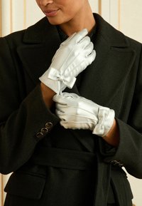 Sudraba satīna cimdi ar krokām un mezgliņiem, ko valkā cilvēks melnā mētelī. Spīdīga tekstūra un izteiksmīgi dekoratīvi akcenti.