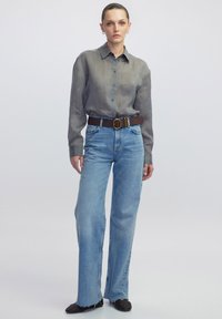 Camicia a maniche lunghe grigia con bottoni, jeans a gamba larga azzurri, cintura marrone con fibbia rotonda, scarpe nere testurizzate, sfondo neutro.