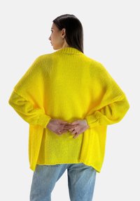 Żółty dziergany sweter o luźnym kroju, z prążkowymi mankietami i wysokim kołnierzem. Tekstura jest miękka i gruba, z rozcięciem na dole.