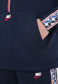 Person som bär en marinblå Nike-träningsoverall med röda, vita och blåa märkestripes på ärmar och ben, med handen i huvan ficka.