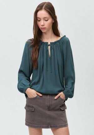 Blusa lunga teal con scollatura a chiave e dettagli con cravatta. Abbinata a una gonna di jeans marrone con tasche laterali. Texture liscia.