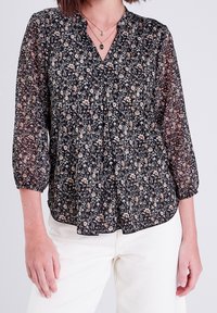 Blouse noire à motifs floraux avec des manches trois-quarts ; tissu léger et transparent présentant des motifs floraux complexes dans des tons crème et pêche.