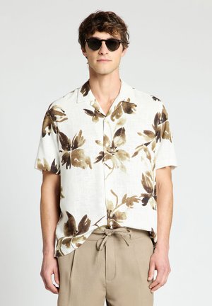 Mann trägt schwarze Sonnenbrille, weißes Kurzarmhemd mit braunem Blumenmuster und beige Kordelzughose vor einfarbigem Hintergrund.