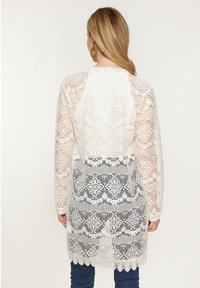 Cardigan lungo in pizzo bianco, caratterizzato da motivi intricati e bordi scallop. Tessuto trasparente con dettagli elaborati, indossato sopra jeans blu.