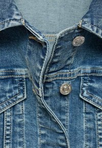 Gros plan sur le col et la partie supérieure de la poitrine d'une veste en denim bleu avec boutons-pression métalliques et poches à rabat doublement cousues.