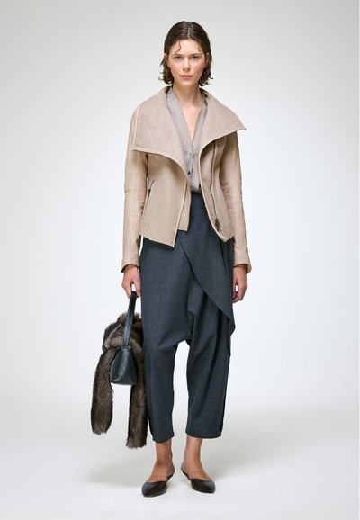 Femme portant une veste beige, un pantalon gris asymétrique, tenant un sac à main noir avec un accessoire en fourrure, debout devant un fond clair uni.