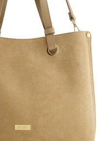 Beżowa torebka wykonana z zamszu o gładkiej teksturze, z złotymi akcentami hardware i podwójnymi ramiączkami na ramię. Logo umieszczone na złotej tabliczce z przodu.