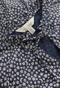 Marinblå bomullsklänning med ett vitt blommönster och himmelska detaljer, volang vid halsringningen och en rosett vid kragen. Lappen är synlig inuti.