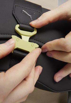 Sac à main noir avec une surface texturée et une boucle en plastique vert olive. Un logo est visible sur la sangle. Des mains manipulent la boucle.