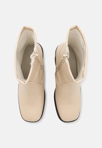 Bottes à chevilles beiges en matériau lisse, avec bout arrondi, semelles noires et fermetures éclair latérales pour un enfilage facile. La tige a une finition douce et mate.